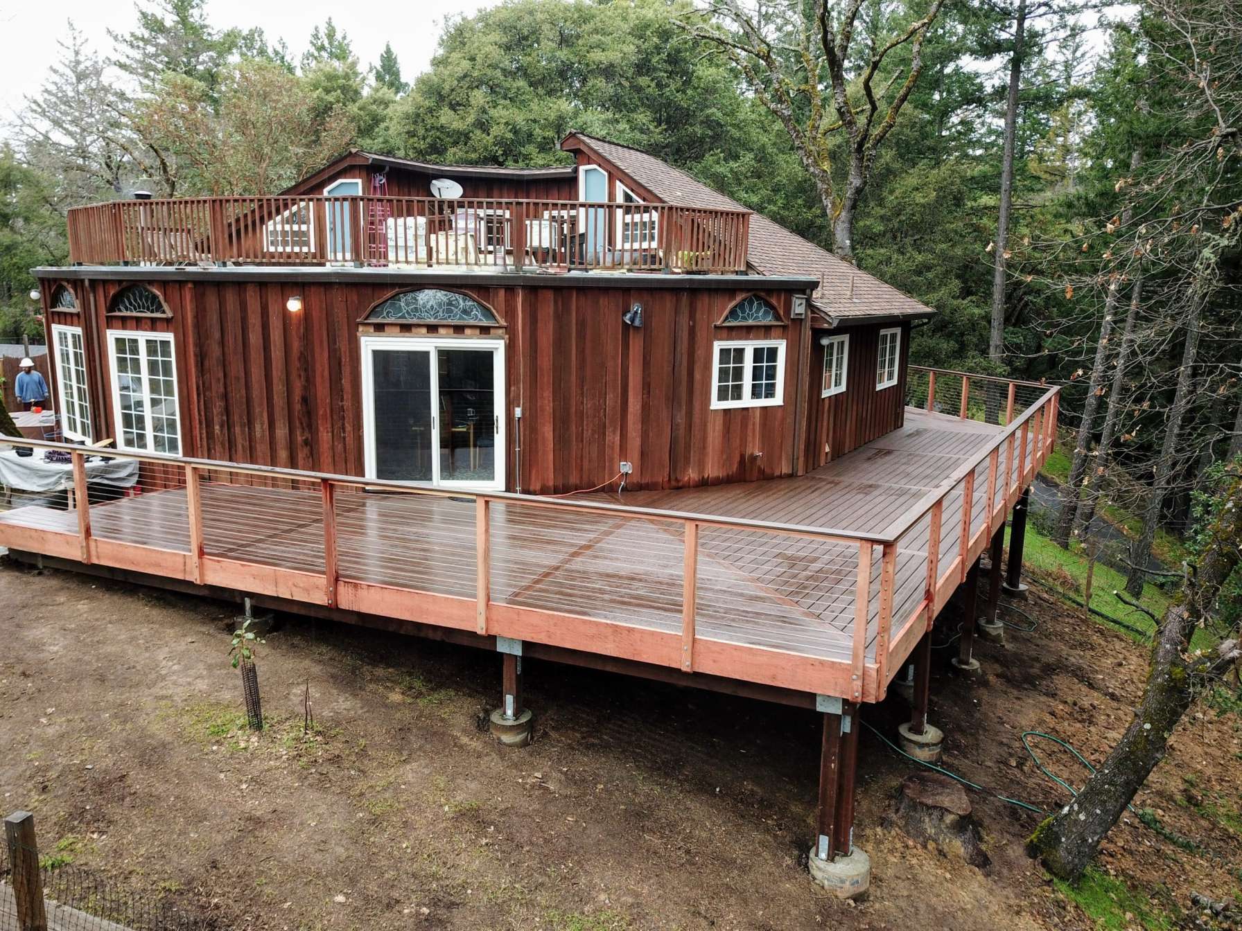 A round wooden house with a large deck, surrounded by trees. The deck features a railing and is elevated above the ground.
